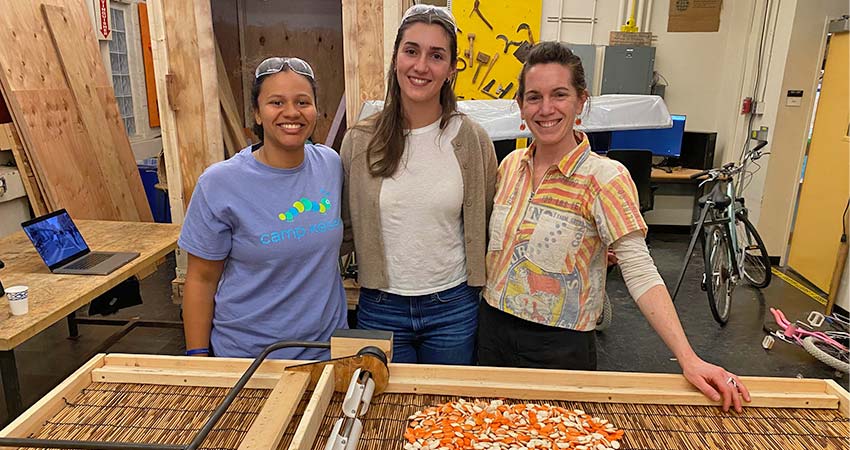 Three smiling woman standing behind a work table.