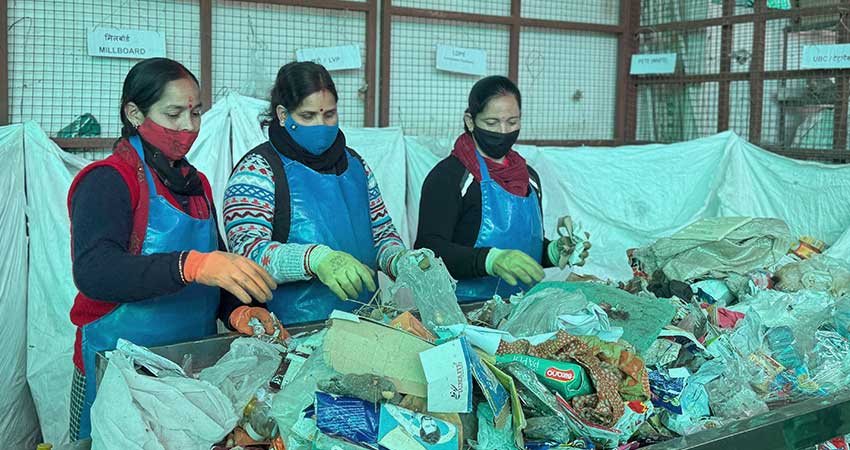 Three women with face masks and gloves sorting garbage on a table.
