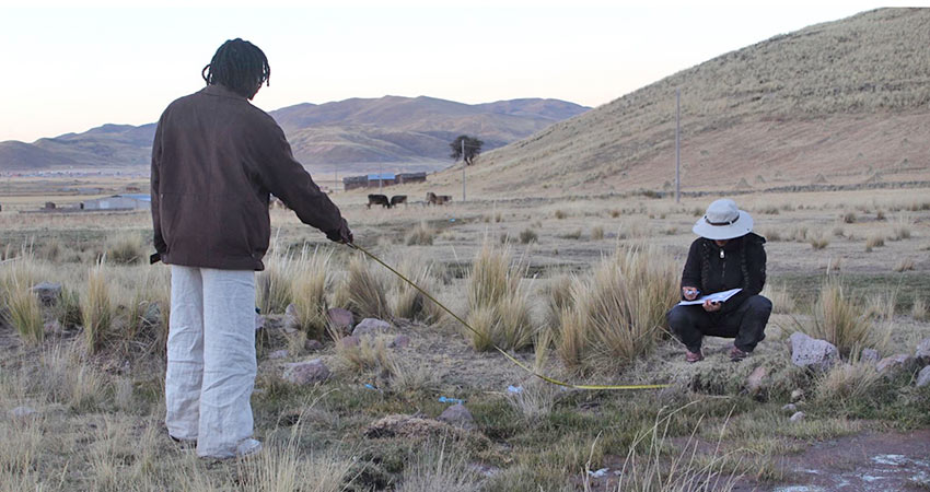 One man standing at left with his back to the camera, another man croucing at right in an arid outdoors site.