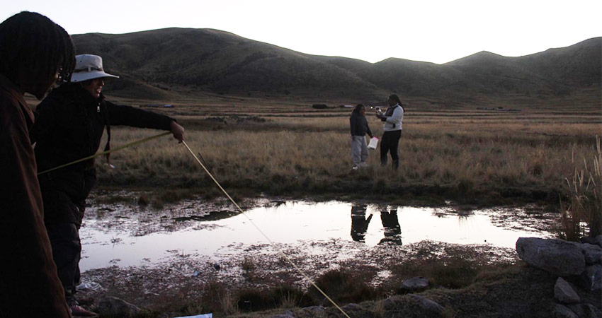 Four people outdoors in dim light on either side of a shallow body of water, mountains in the distance.