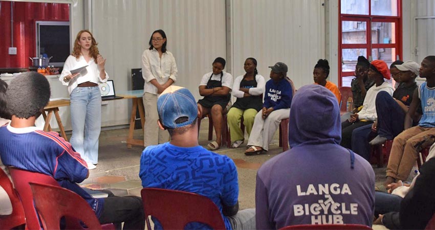 A dozen are so people seated in chairs in a semicircle facing two standing women, one of whom is speaking.