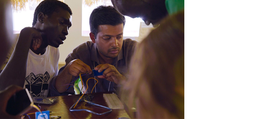 A man sitting at a table holding a couple of wires while other men look on.